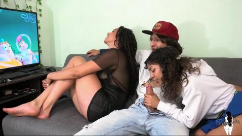 Um cara está assistindo TV com duas garotas, e uma delas está fazendo um boquete.